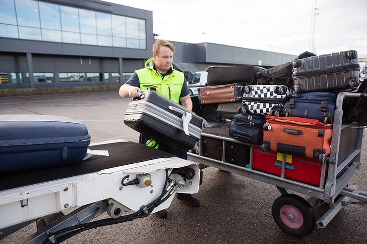 Man in neon vest loads a suitcase onto a luggage cart stacked with bags outside an airport terminal.