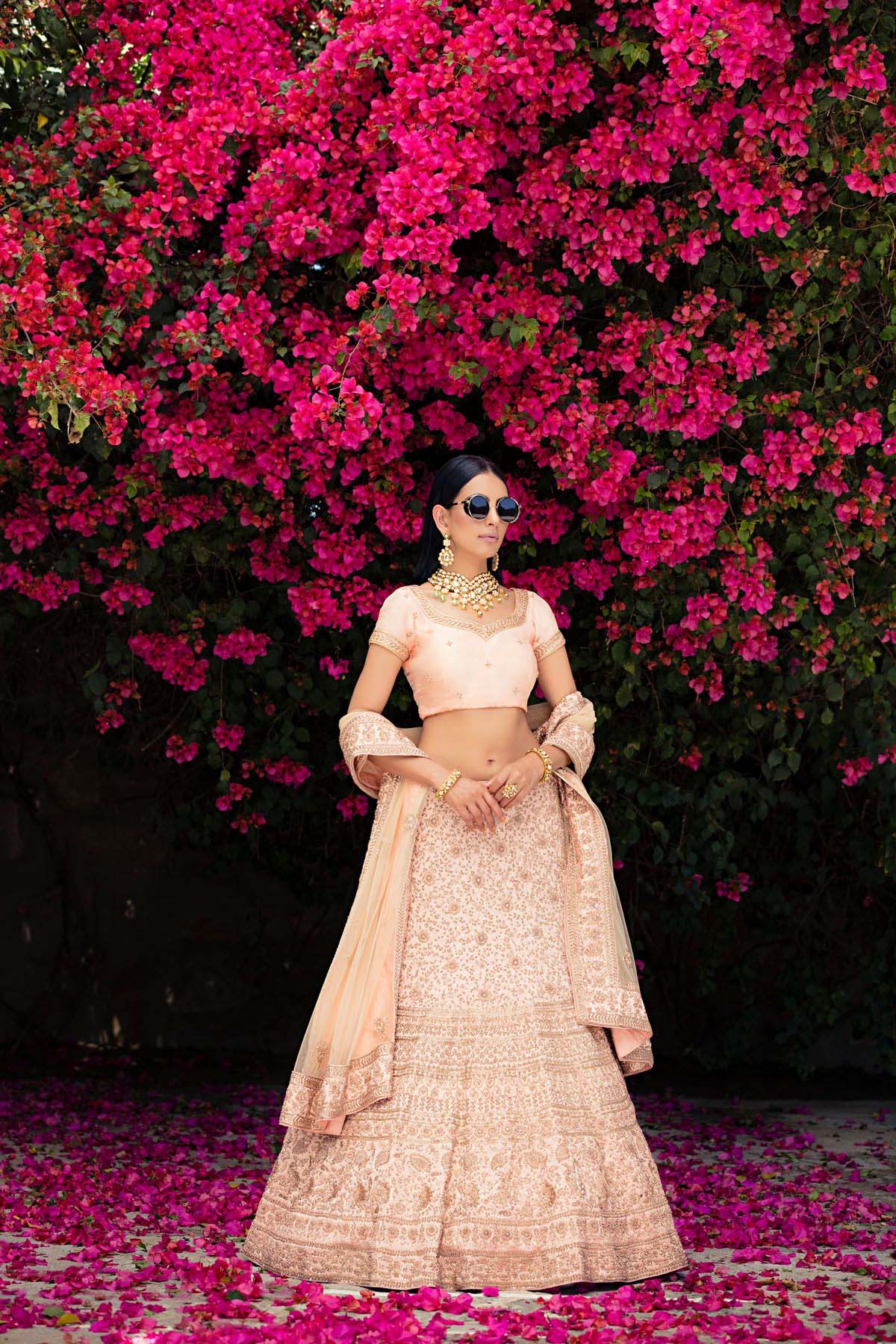 Modern Indian bride standing confidently in front of a backdrop of pink flowers,wearing a pastel peach lehenga and gold jewelry.