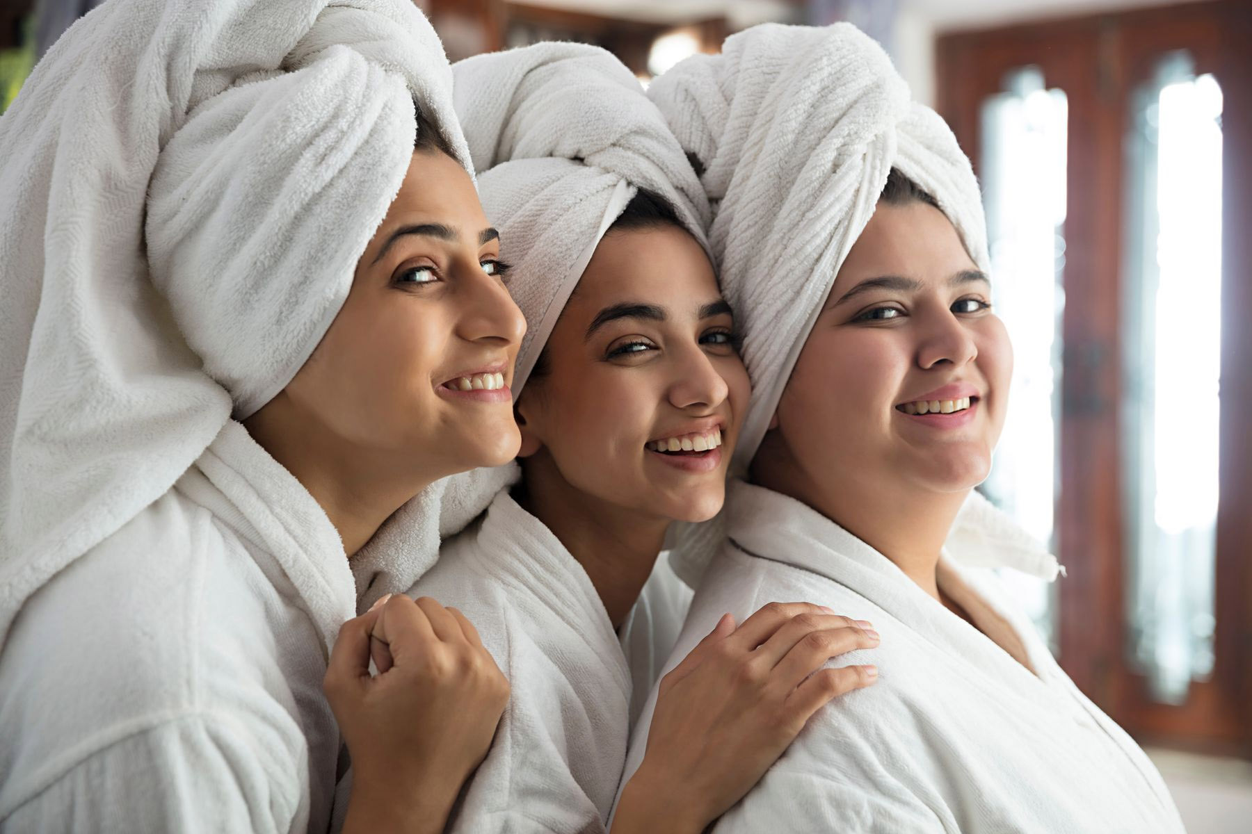 Three smiling women in white bathrobes with towels wrapped on their heads, enjoying a pre-bridal relaxation session.