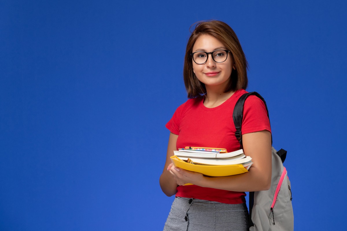 front-view-female-student-red-shirt-with-backpack-holding-books-files-smiling-blue-wall (1).jpg